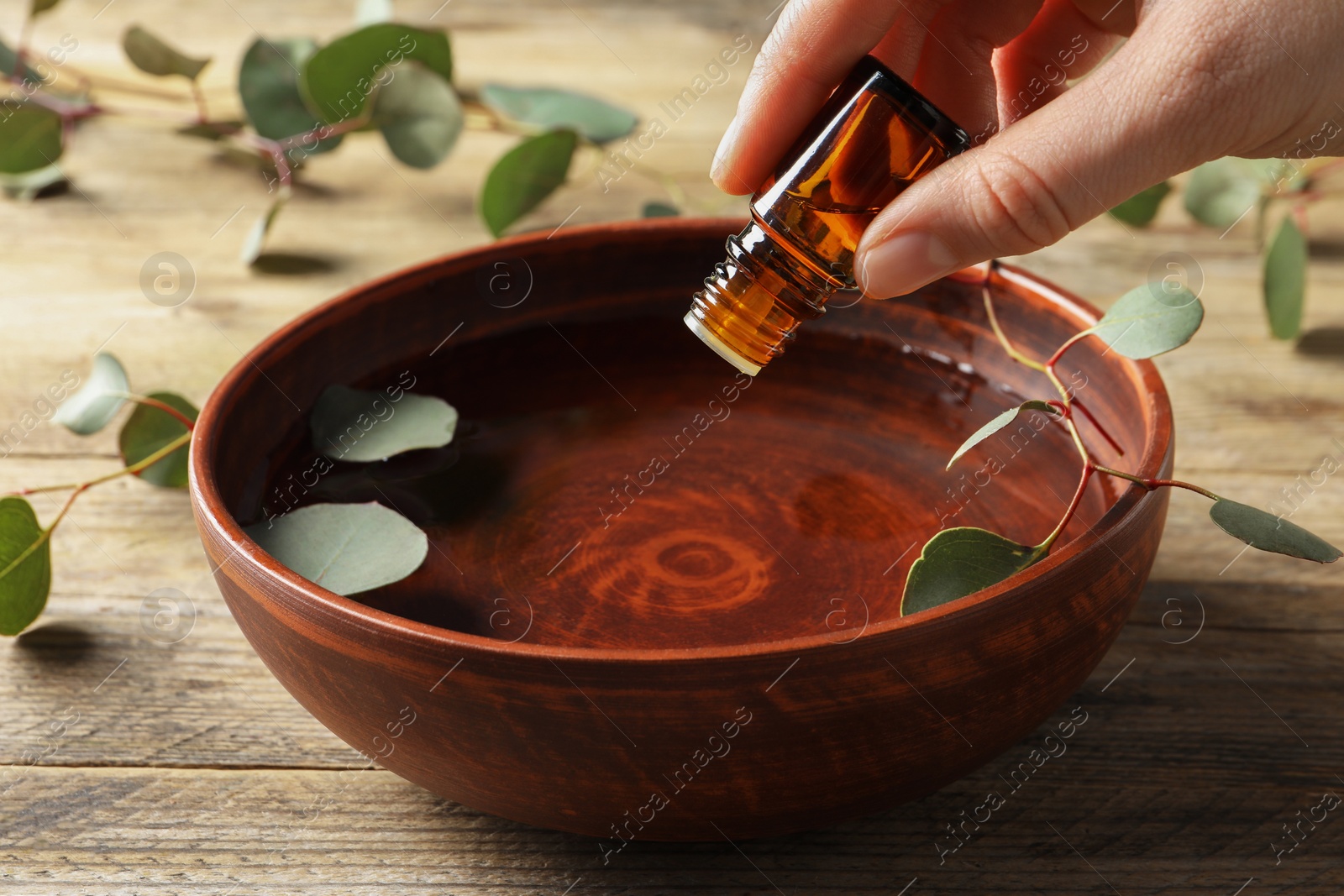 Photo of Woman dripping eucalyptus essential oil from bottle into bowl at wooden table, closeup