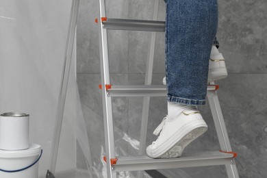 Woman climbing metallic folding ladder, can and bucket of paint near grey wall, closeup