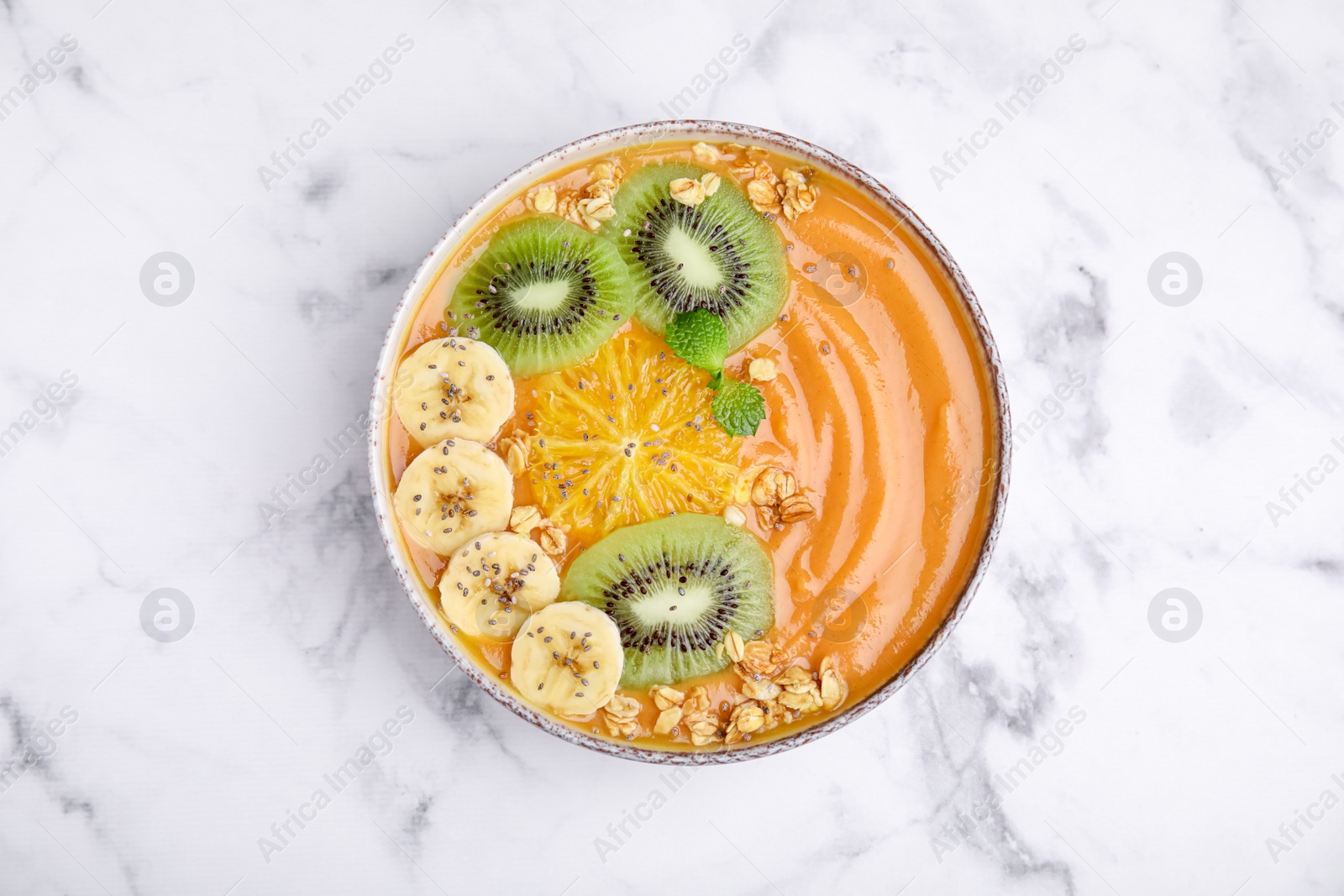 Photo of Bowl of delicious fruit smoothie with fresh banana, kiwi slices and granola on white marble table, top view