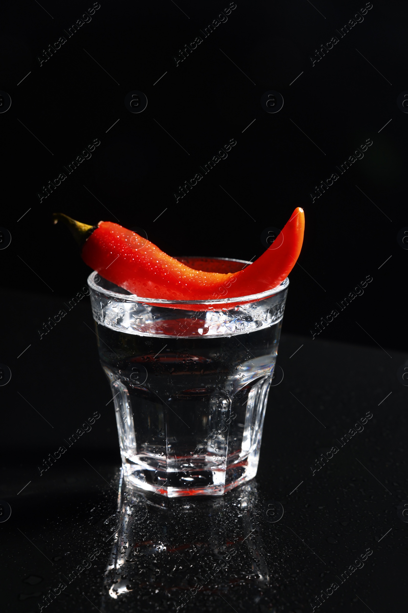 Photo of Red hot chili pepper and vodka in glass on black table