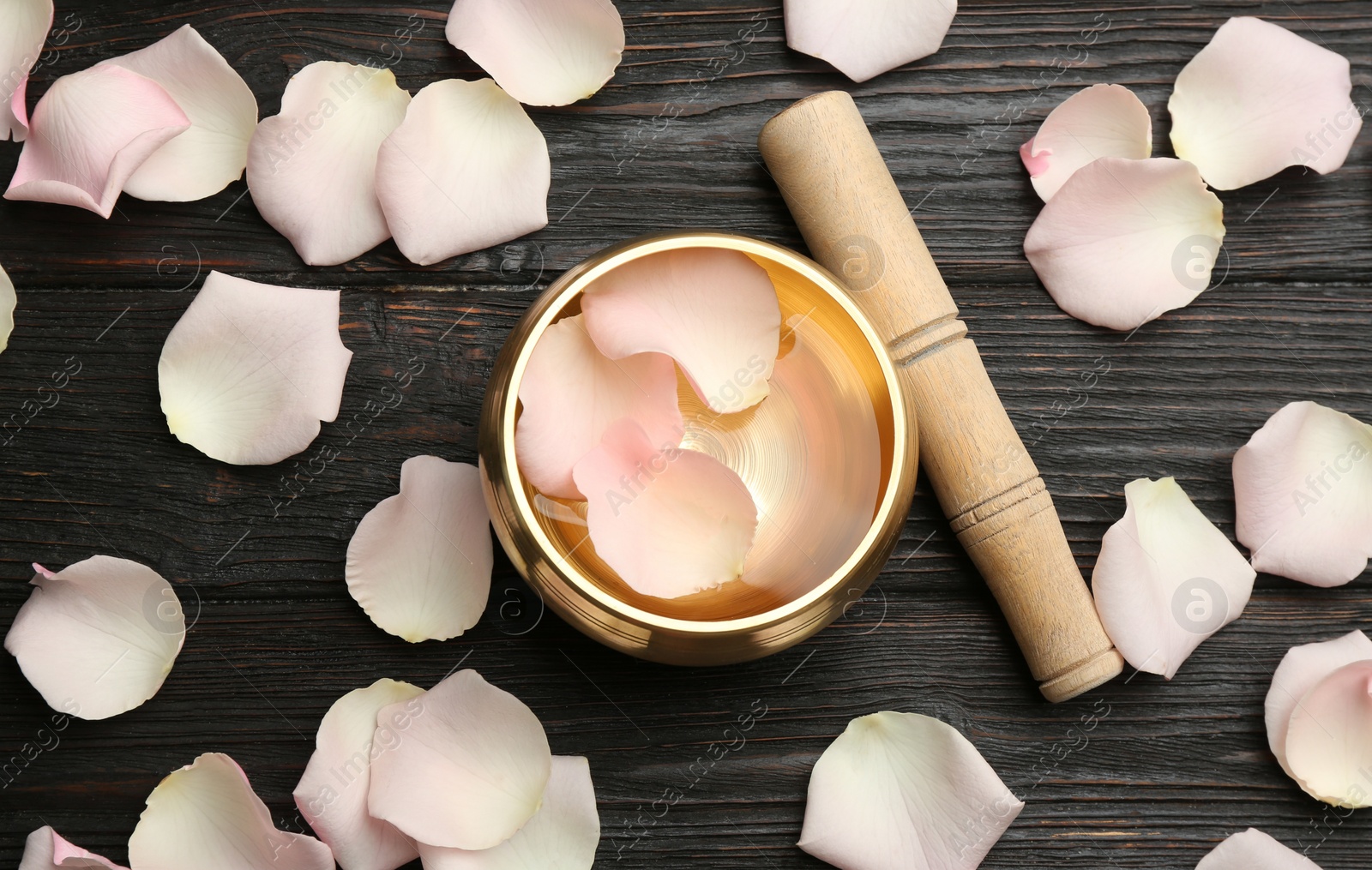 Photo of Golden singing bowl, mallet and petals on black wooden table, flat lay. Sound healing