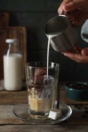 Photo of Woman pouring milk into glass with coffee ice cubes at wooden table, closeup