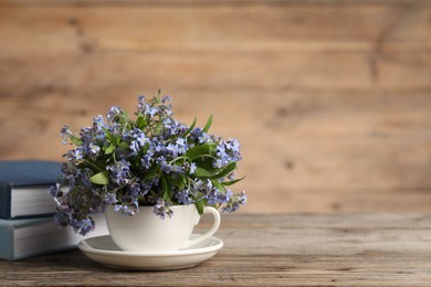 Photo of Beautiful forget-me-not flowers in cup, saucer and books on wooden table, closeup. Space for text