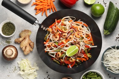 Shrimp stir fry with noodles and vegetables in wok surrounded by ingredients on grey table, flat lay