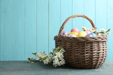 Photo of Painted Easter eggs in wicker basket and blossoming branches on table against color background. Space for text