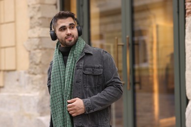 Photo of Handsome man in warm scarf and headphones near building outdoors. Space for text