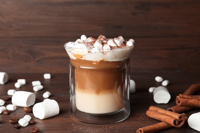 Photo of Glass with delicious coffee drink, marshmallows and cinnamon sticks on wooden table