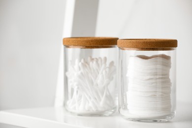 Photo of Glass jars with cotton pads and swabs on white shelf, closeup. Space for text