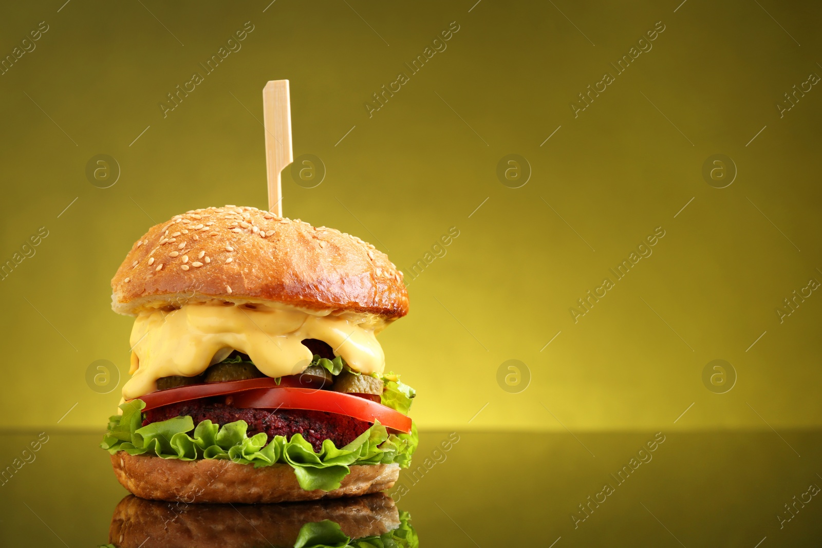 Photo of Delicious vegetarian burger on mirror surface against olive background, space for text