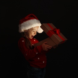 Photo of Cute little girl in Santa hat with Christmas gift box on black background