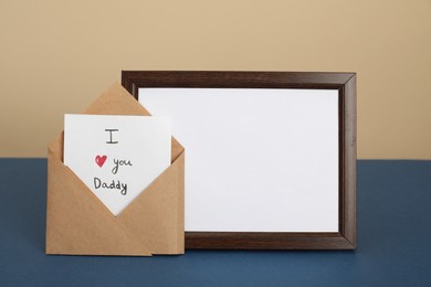 Photo of Happy Father's Day. Card with phrase I Love You, Daddy in envelope and empty frame on blue table, closeup. Space for text