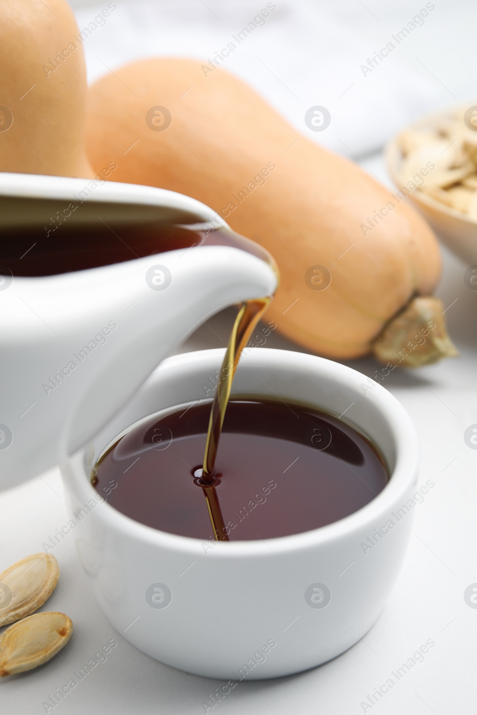 Photo of Pouring pumpkin seed oil into bowl on white table, closeup