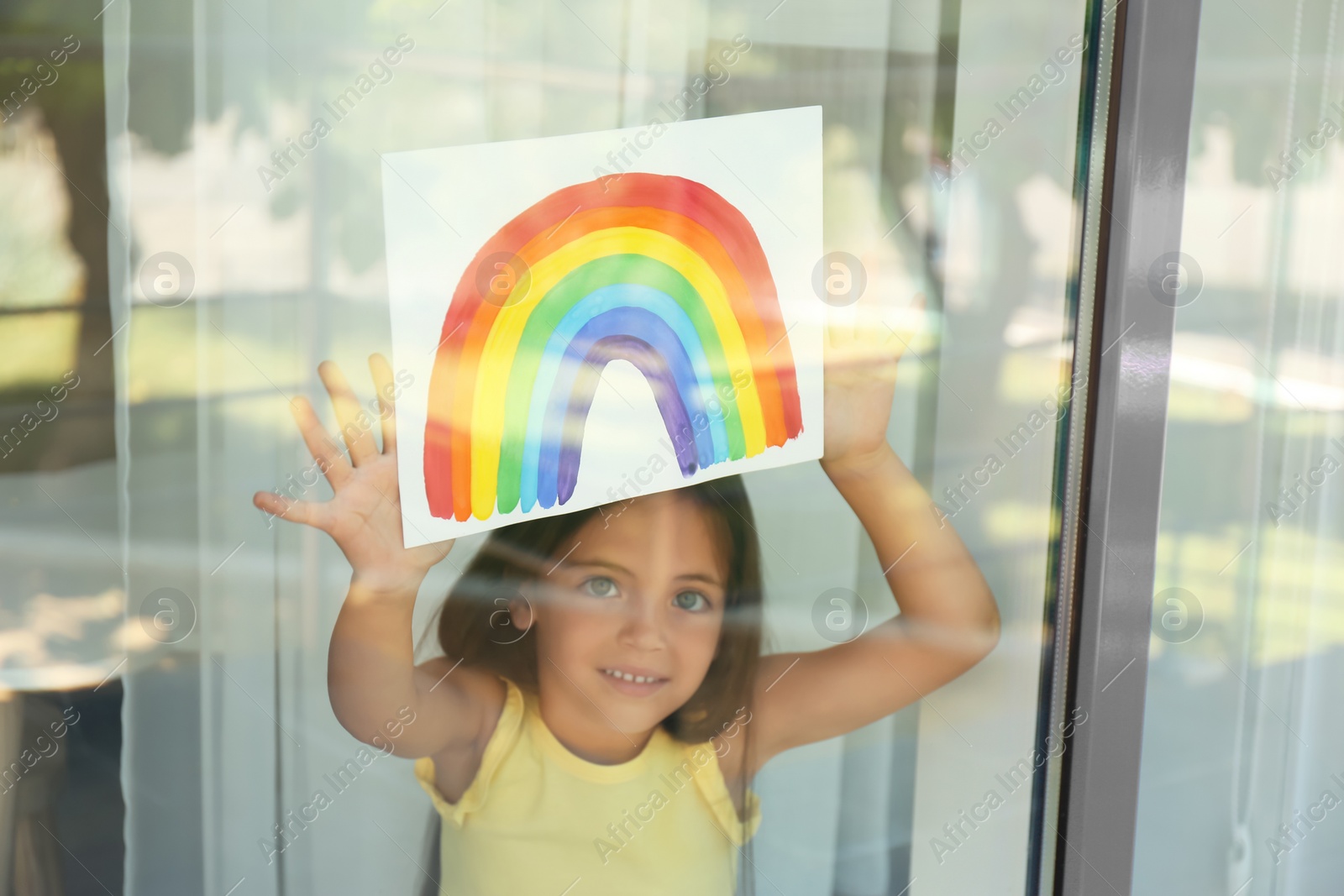 Photo of Little girl with picture of rainbow near window, view from outdoors. Stay at home concept