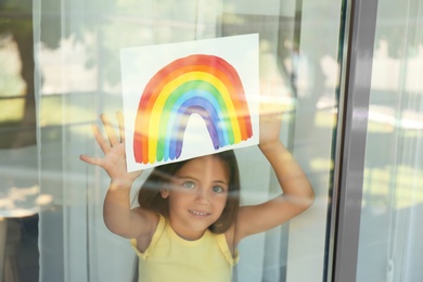 Little girl with picture of rainbow near window, view from outdoors. Stay at home concept