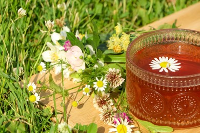 Photo of Ornate glass cup of tea, different wildflowers and herbs on wooden board in meadow, closeup