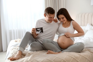 Young pregnant woman and her husband with ultrasound picture of baby in bedroom