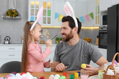 Photo of Father and his cute daughter having fun while painting Easter eggs at table in kitchen