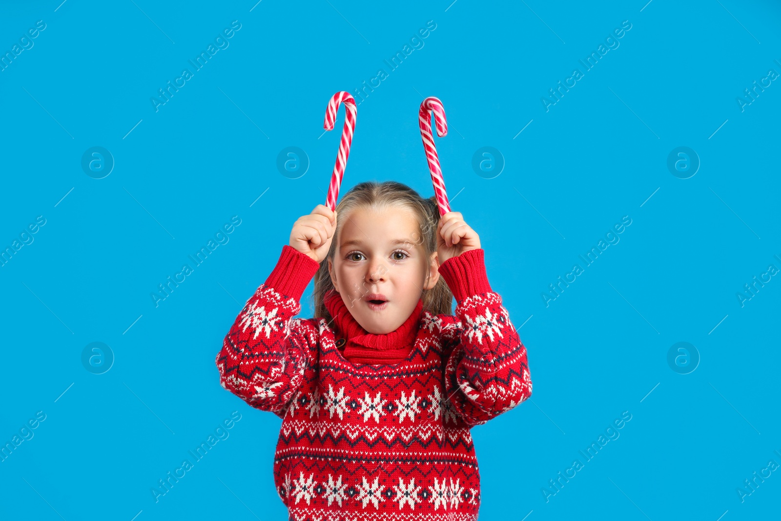 Photo of Surprised little girl in knitted Christmas sweater holding candy canes on blue background