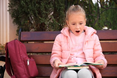 Photo of Cute little girl with backpack reading book on bench outdoors