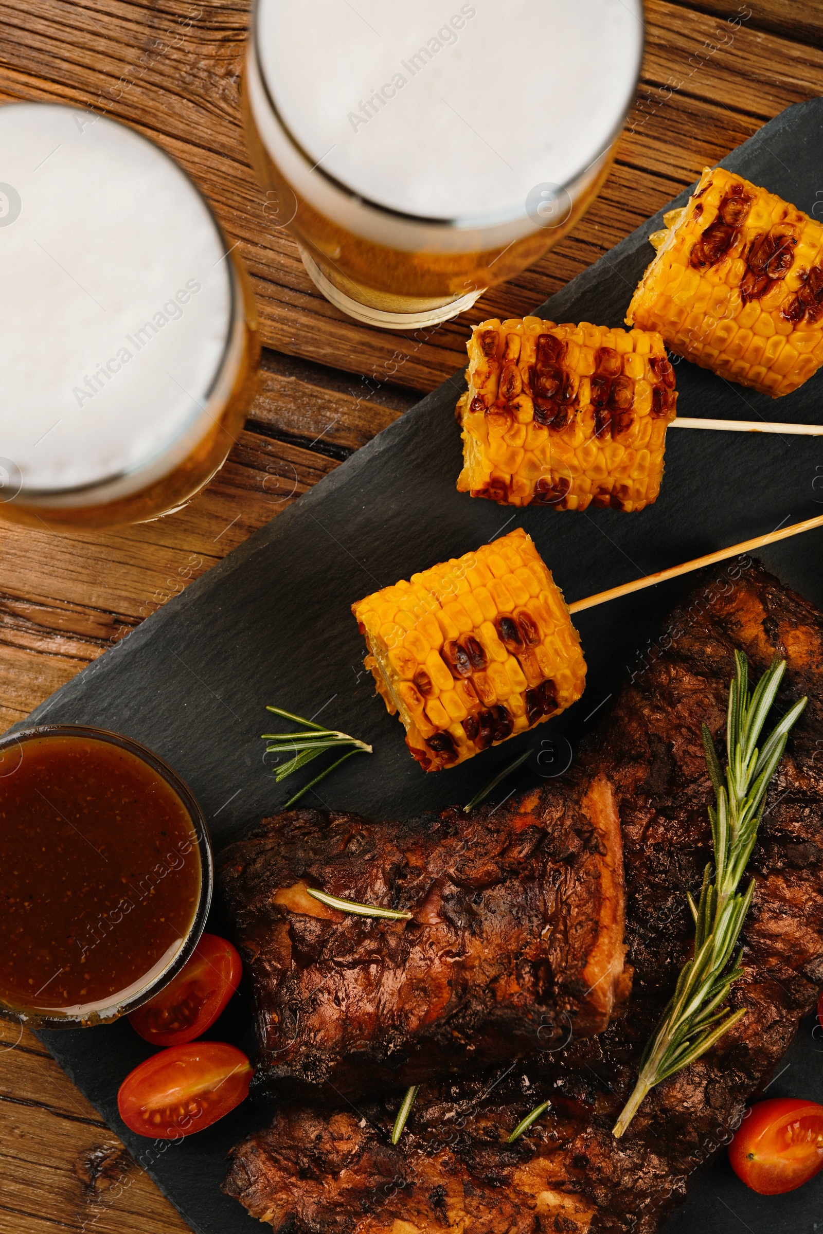 Photo of Glasses of beer, delicious grilled ribs, corn and sauces on wooden table, flat lay