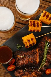 Glasses of beer, delicious grilled ribs, corn and sauces on wooden table, flat lay