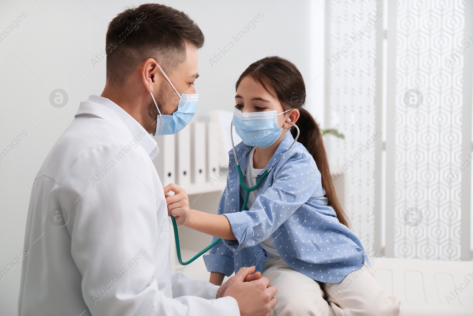 Photo of Little girl playing with pediatrician during visit in hospital. Doctor and patient wearing protective masks