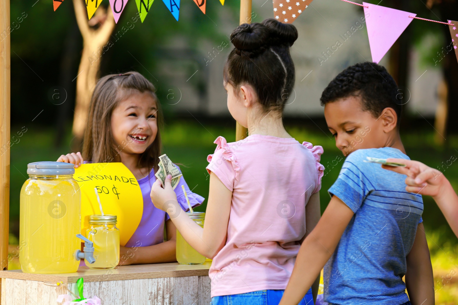 Photo of Little girl selling natural lemonade to kids in park. Summer refreshing drink