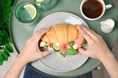 Photo of Woman holding tasty croissant sandwich over plate at table, top view