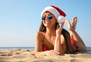 Photo of Beautiful young woman in Santa hat and bikini on beach, space for text. Christmas vacation