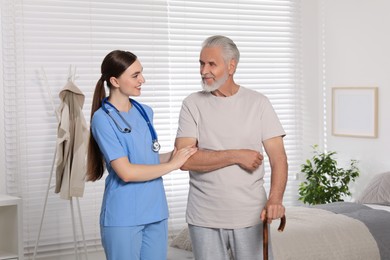 Photo of Young healthcare worker assisting senior man with walking cane indoors