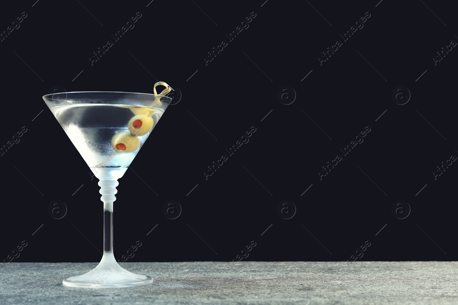 Photo of Martini cocktail with olives on grey table against dark background, space for text