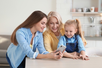 Photo of Portrait of young woman, her mother and daughter with smartphone at table indoors