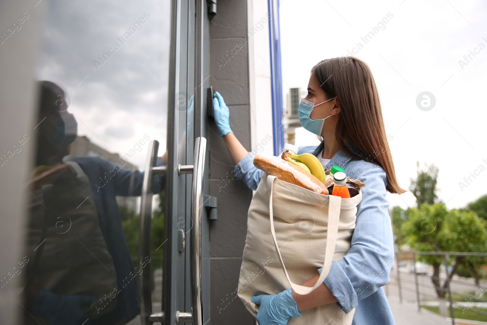 Photo of Female volunteer with products ringing doorbell outdoors. Aid during coronavirus quarantine