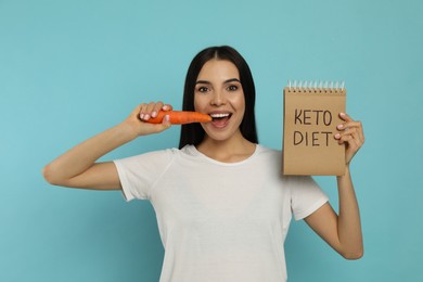 Happy woman eating carrot and holding notebook with words Keto Diet on turquoise background