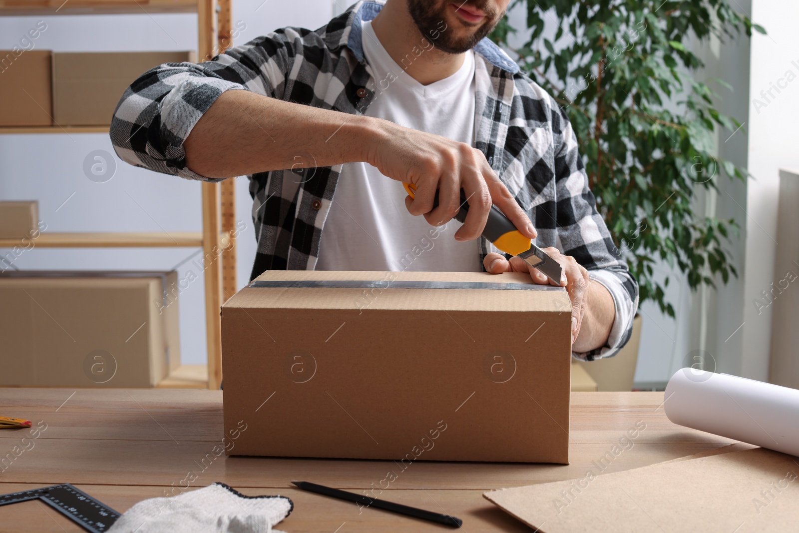 Photo of Man using utility knife to open parcel at wooden table indoors, closeup