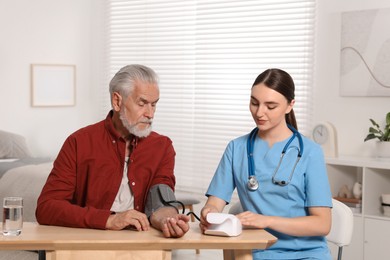 Young healthcare worker measuring senior man's blood pressure at wooden table indoors