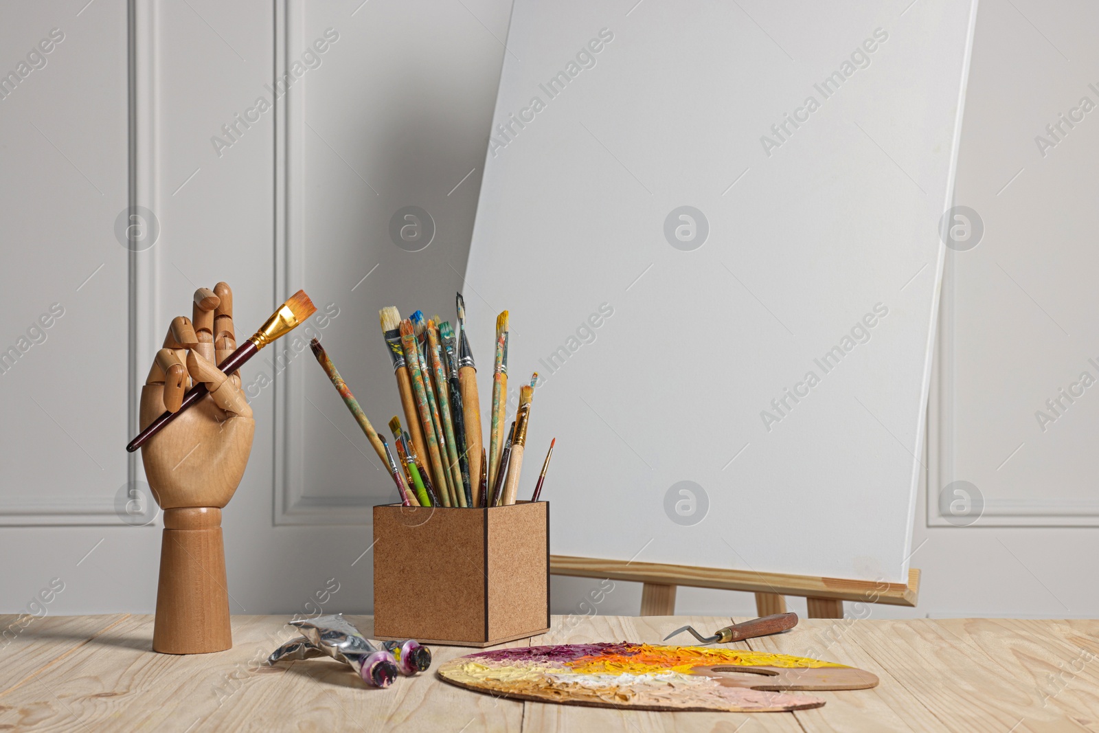 Photo of Easel with blank canvas, hand model and different art supplies on wooden table near white wall