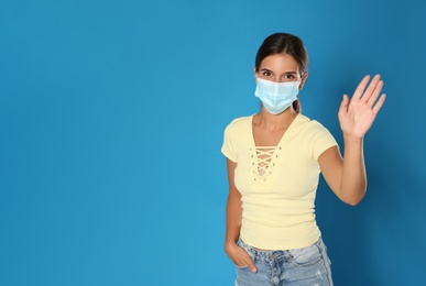 Photo of Woman in protective mask showing hello gesture on blue background, space for text. Keeping social distance during coronavirus pandemic