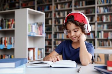 Cute little boy with headphones reading books at table in library