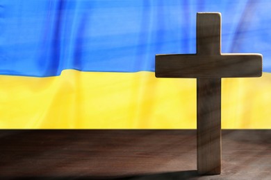 Image of Pray for Ukraine. Cross on wooden table against Ukrainian flag, space for text