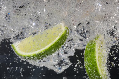 Photo of Juicy lime slices and ice cubes in soda water against black background, closeup