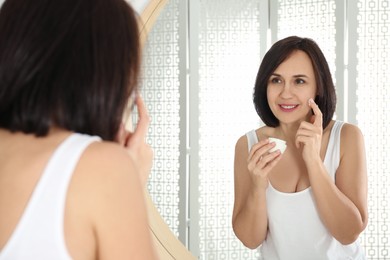 Photo of Happy mature woman applying cream onto face near mirror in bathroom