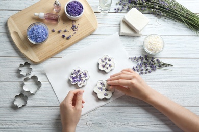 Young woman making handmade soap bar with lavender flowers on white wooden background, top view