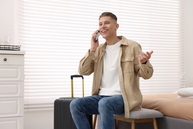 Photo of Smiling guest with suitcase talking on smartphone in stylish hotel room