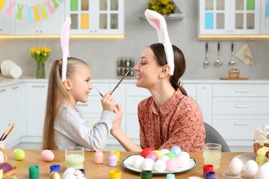 Photo of Mother and her cute daughter having fun while painting Easter eggs at table in kitchen