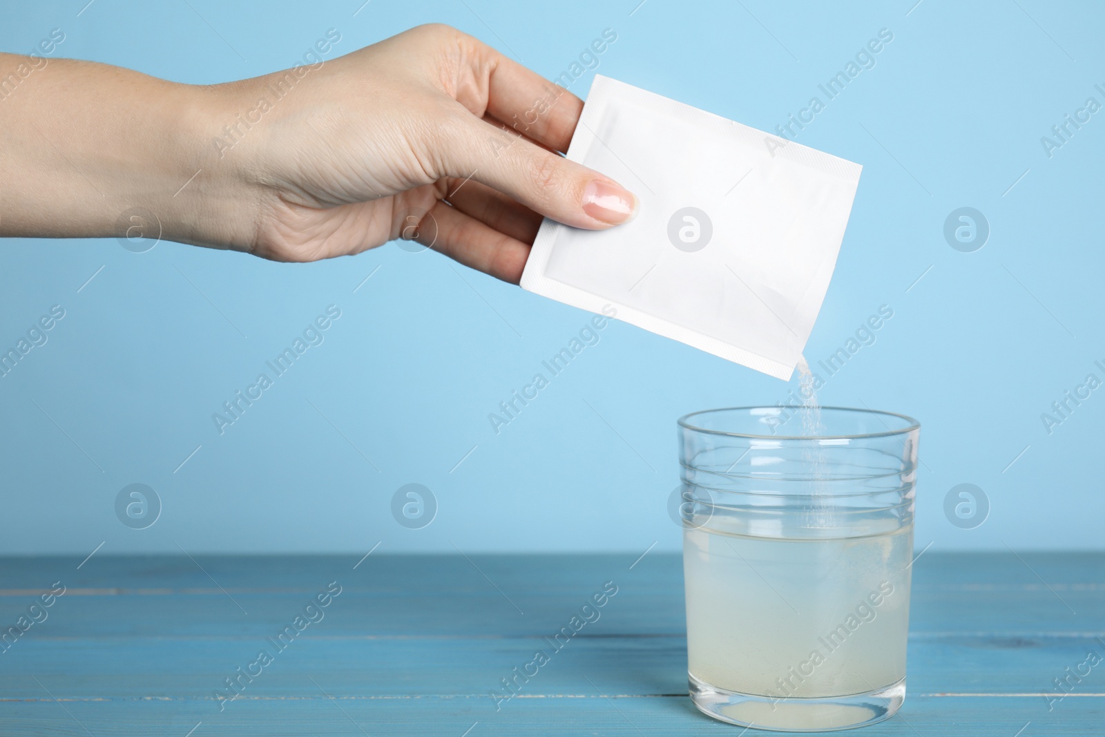 Photo of Woman pouring powder from medicine sachet into glass of water on blue wooden table, closeup