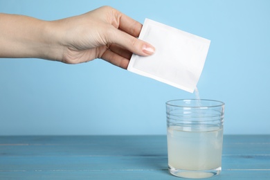 Woman pouring powder from medicine sachet into glass of water on blue wooden table, closeup