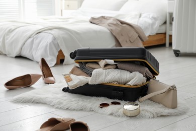 Open suitcase full of clothes, shoes and fashionable accessories on floor in bedroom