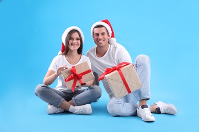 Photo of Beautiful happy couple in Santa hats sitting with Christmas gifts on light blue background