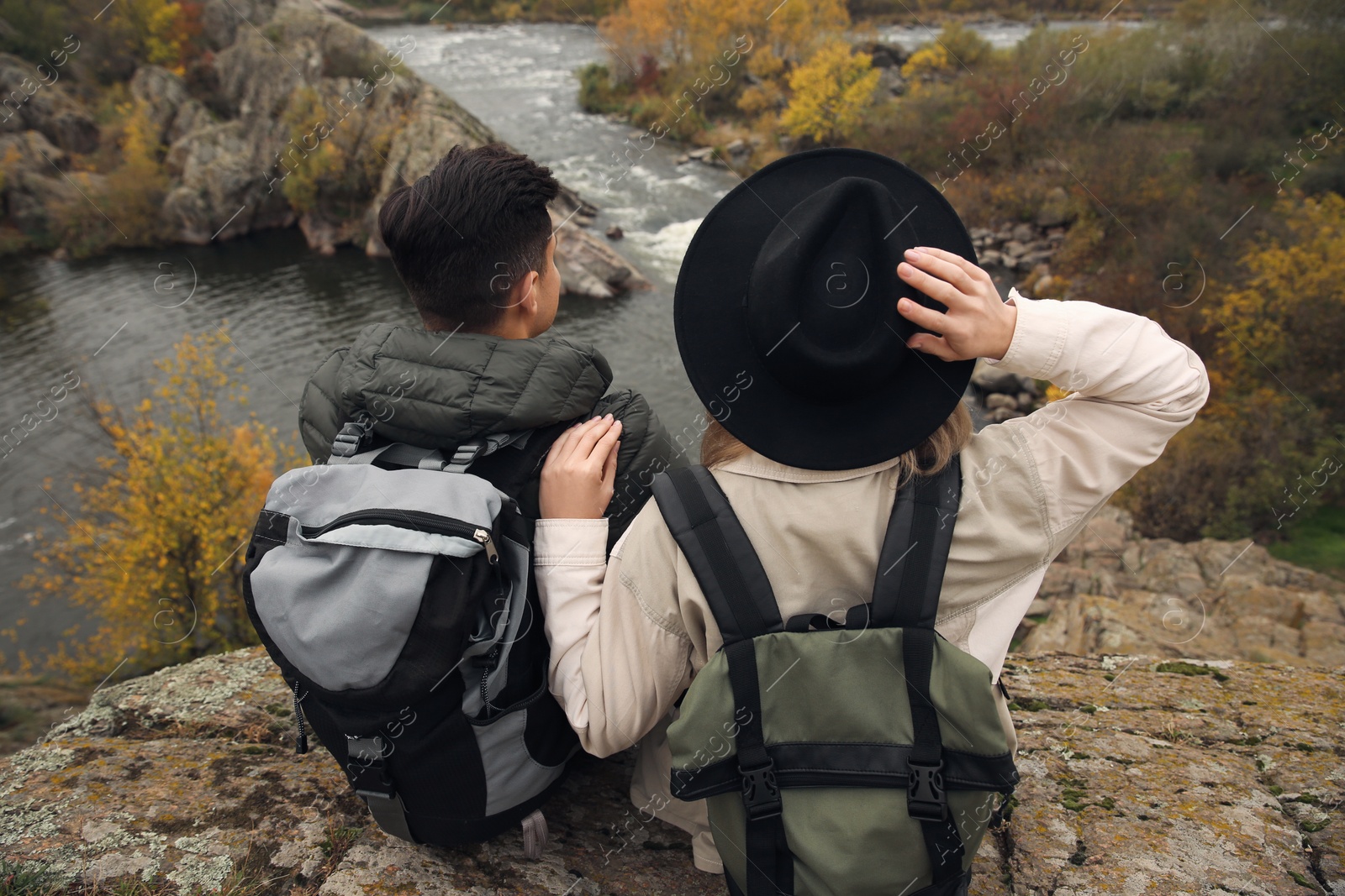 Photo of Couple of hikers with travel backpacks sitting on steep cliff near mountain river, back view